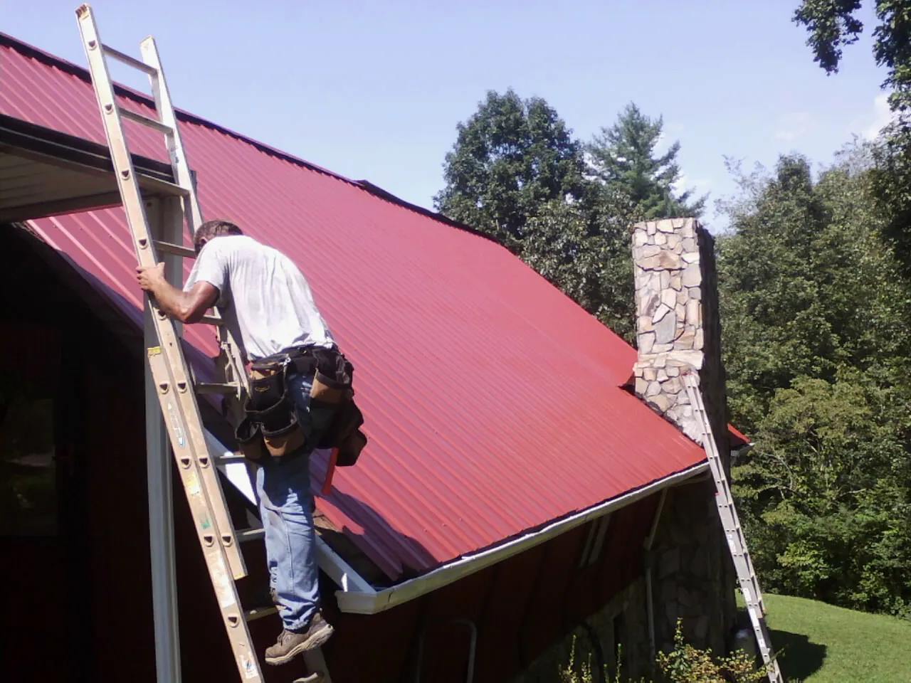 Roofer on a ladder beside a red metal roof and stone chimney — Soffit & Fascia Repair by Roofing Brothers in Fort Bragg